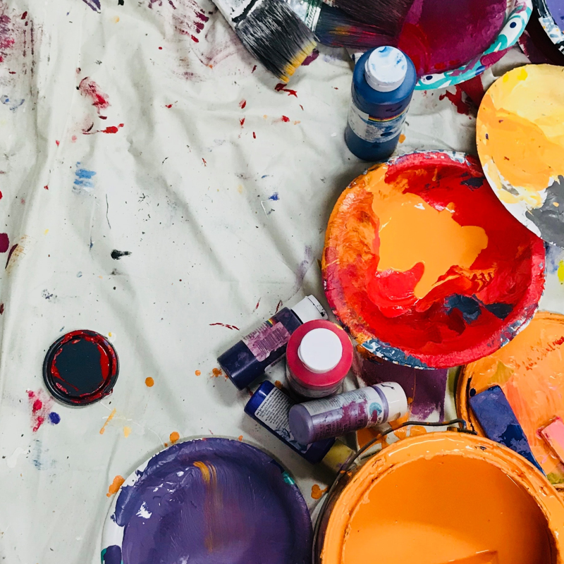 paint bottles, brushes and trays placed on a white sheet with spots of spilled paint.